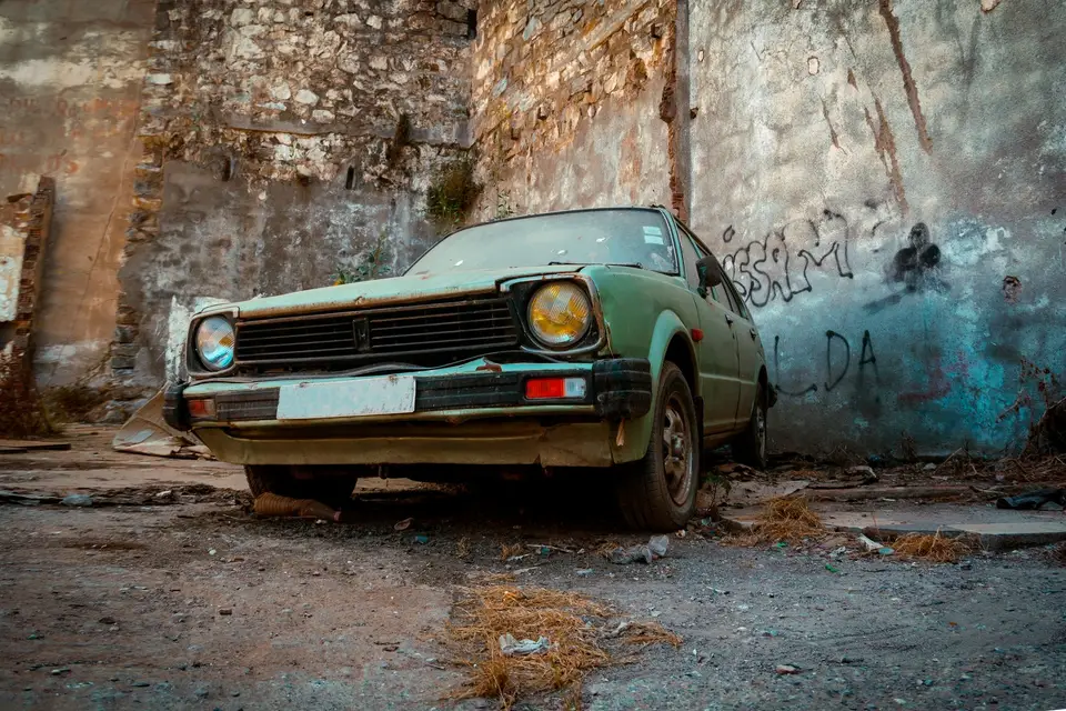 A rusty, faded green hatchback parked in front of a graffiti-covered wall